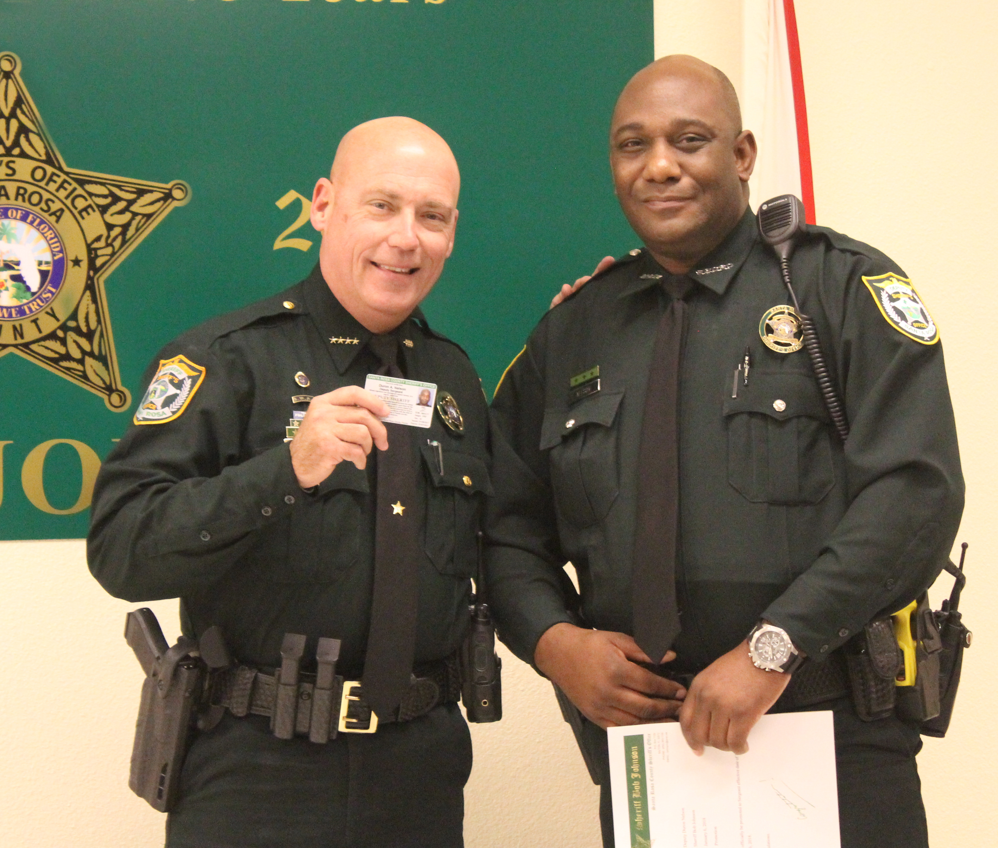 Two police officers, one holding an id card, smiling and standing side by side in a room with a green banner in the background.