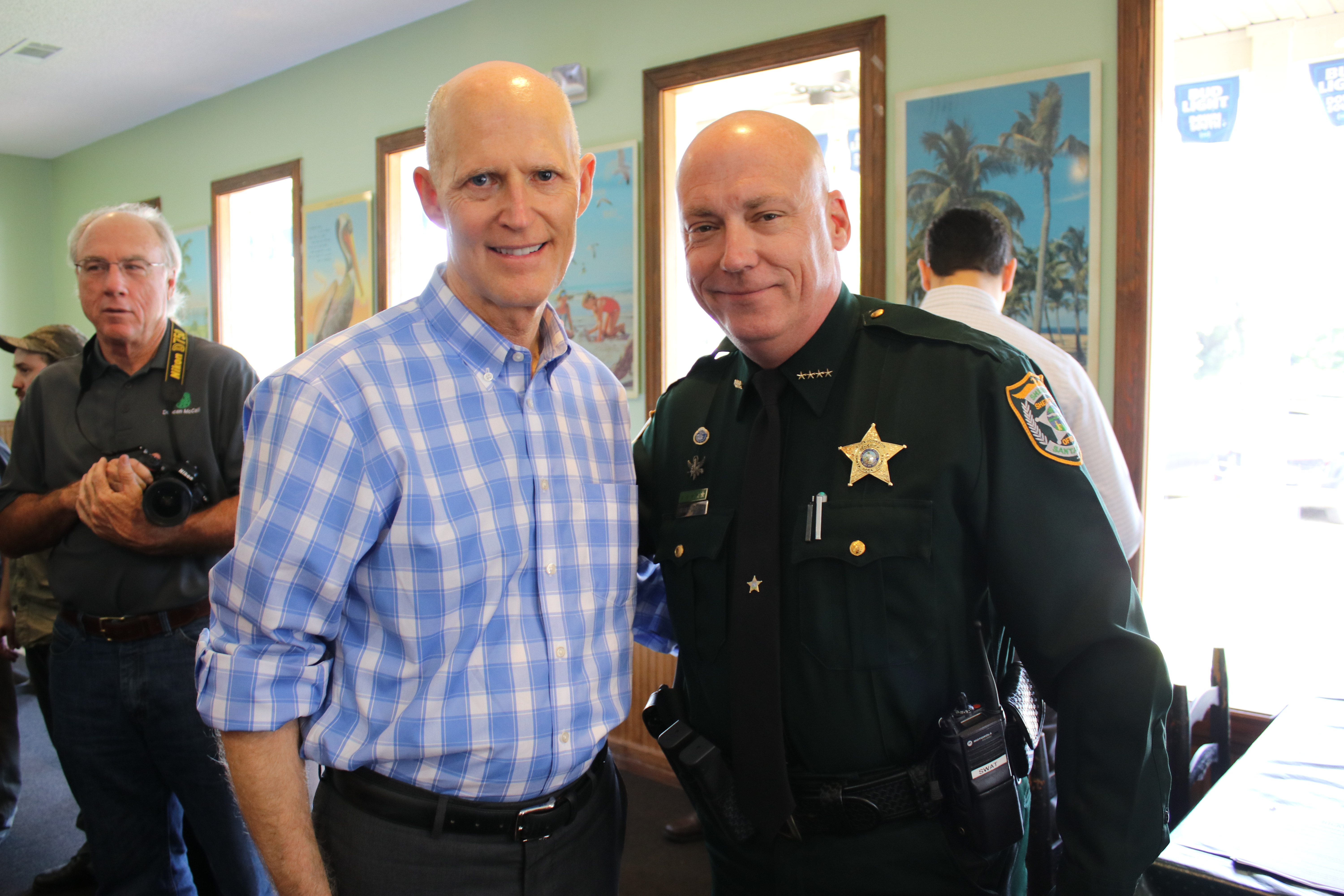 Two men, one in a blue plaid shirt and the other in a green sheriff's uniform, pose together smiling in a room with onlookers.