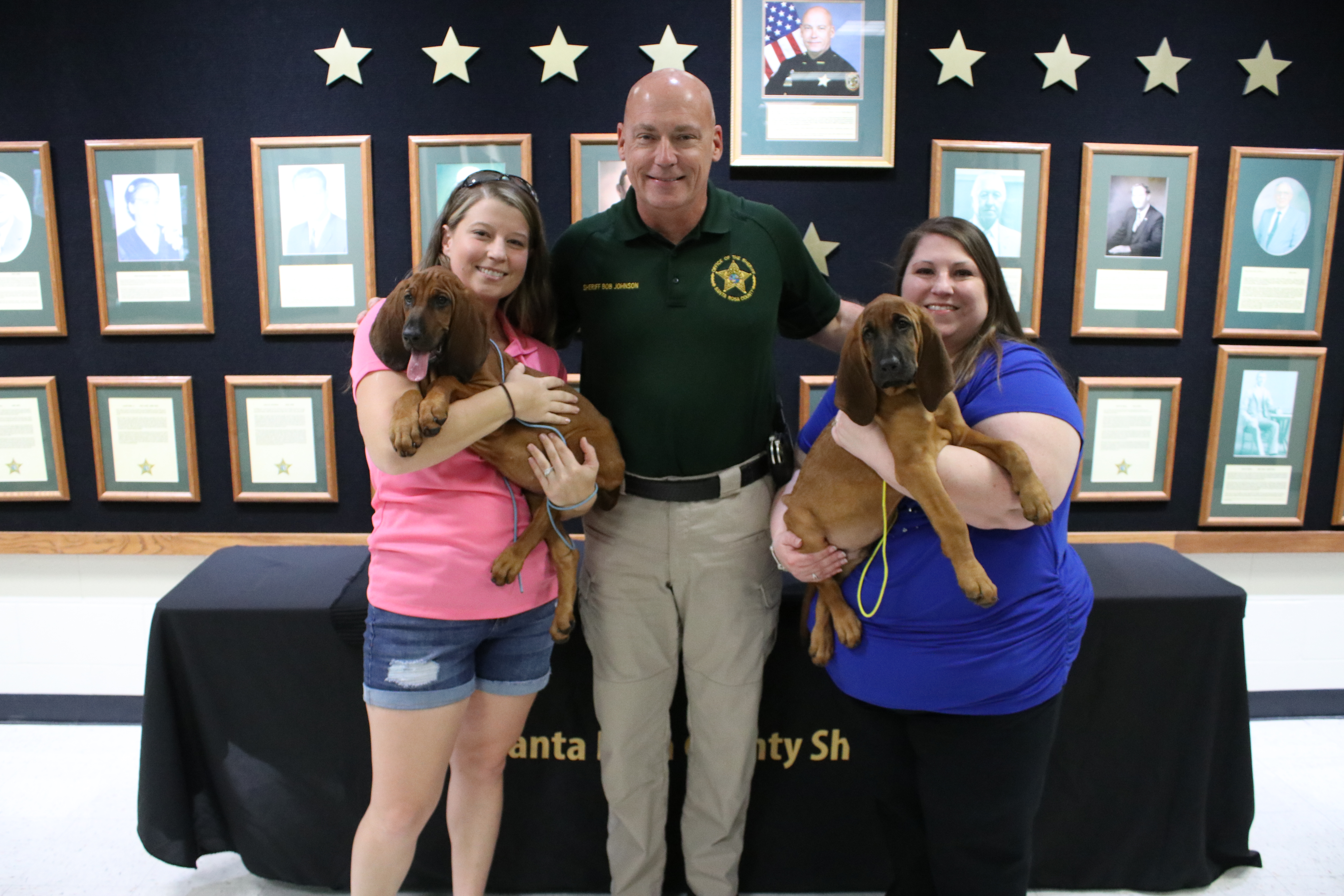 Two women and a sheriff holding puppies stand in front of a wall displaying commemorative plaques and stars.