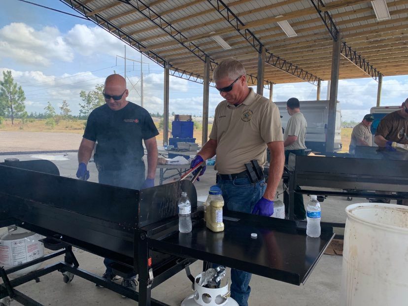 Two men cooking on a large outdoor grill at a community event, with one man flipping food and the other observing.