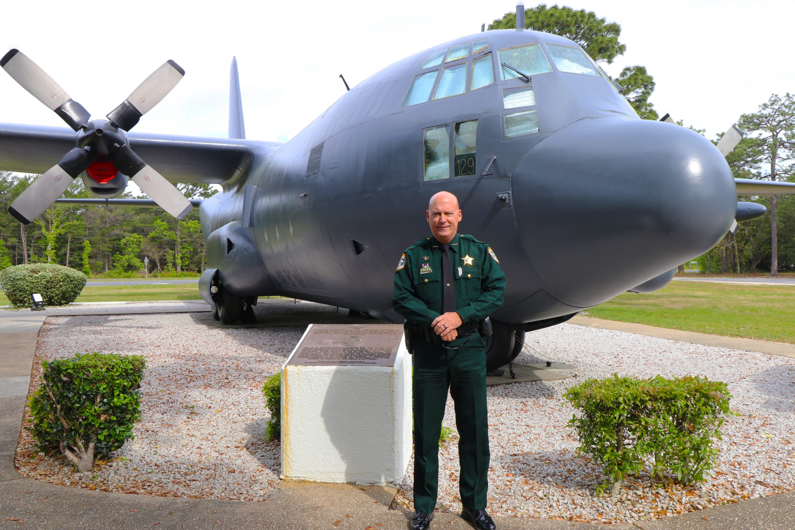 A sheriff in uniform standing in front of a large military transport aircraft with propellers, positioned outdoors.