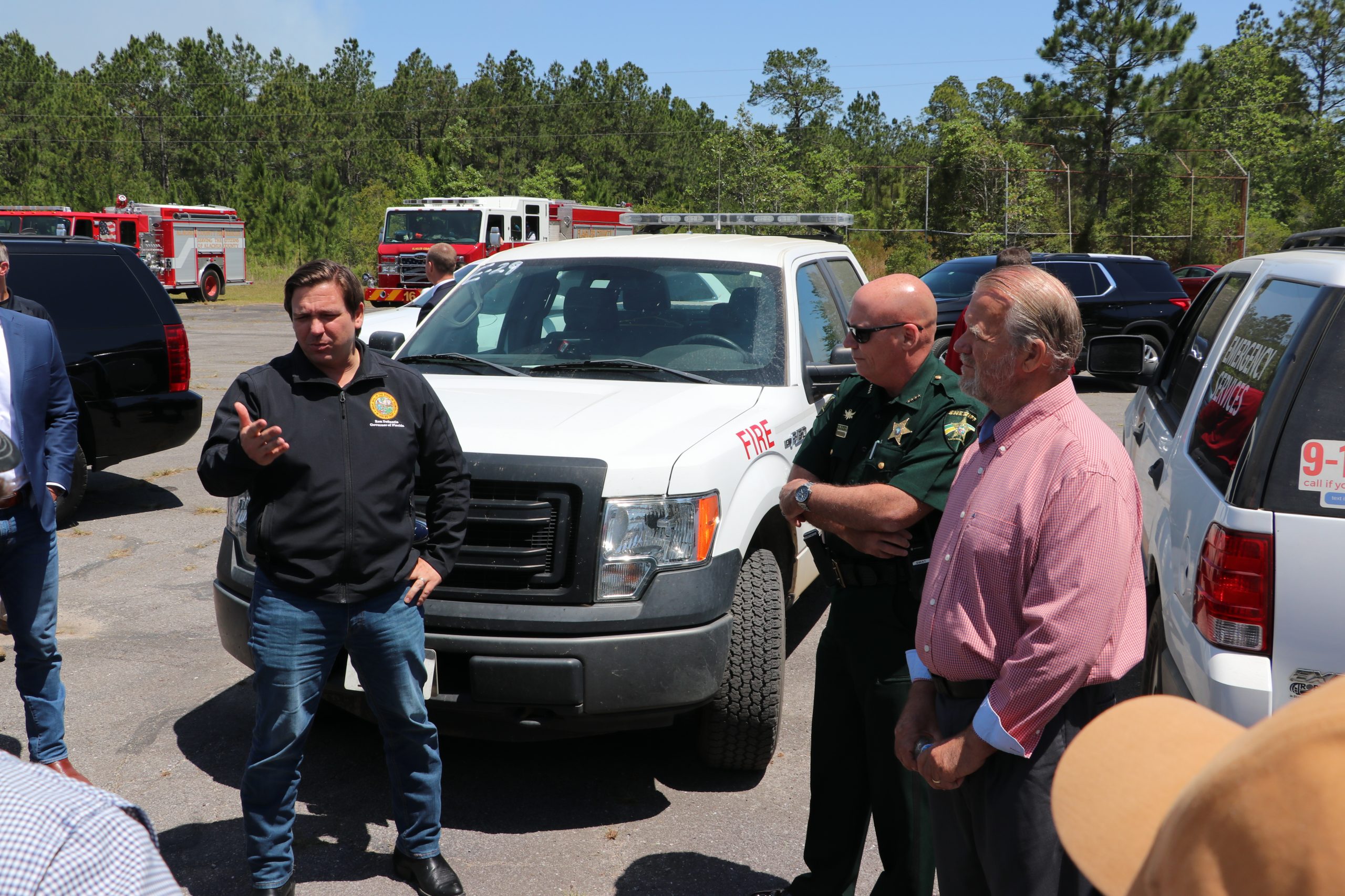 Governor and sheriff discuss with officials in parking lot filled with emergency vehicles under sunny skies.