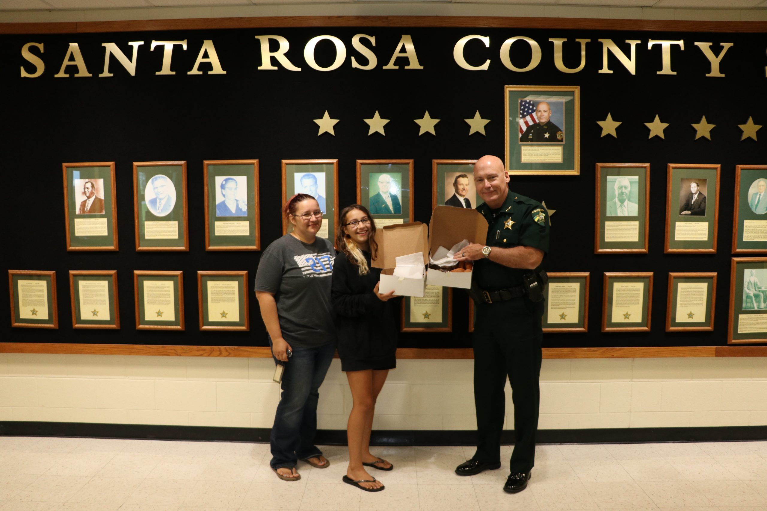 Two women and a sheriff's deputy smiling while standing in front of a wall titled "santa rosa county" adorned with framed portraits and stars.