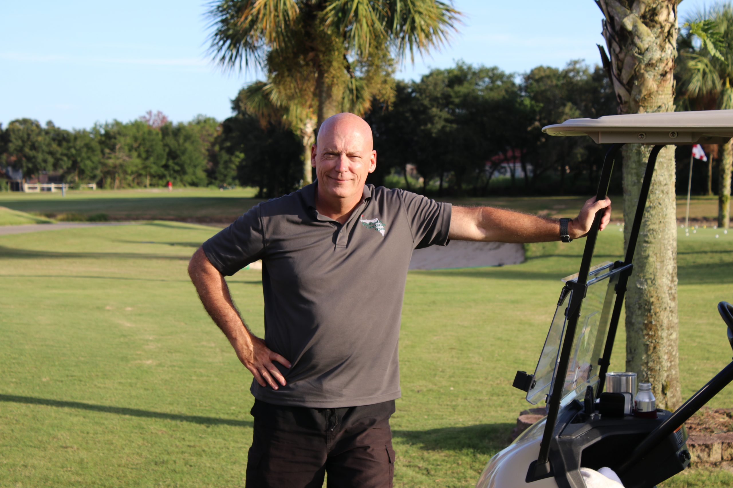 A bald man in a black polo shirt stands smiling beside a golf cart on a sunny golf course, with palm trees in the background.