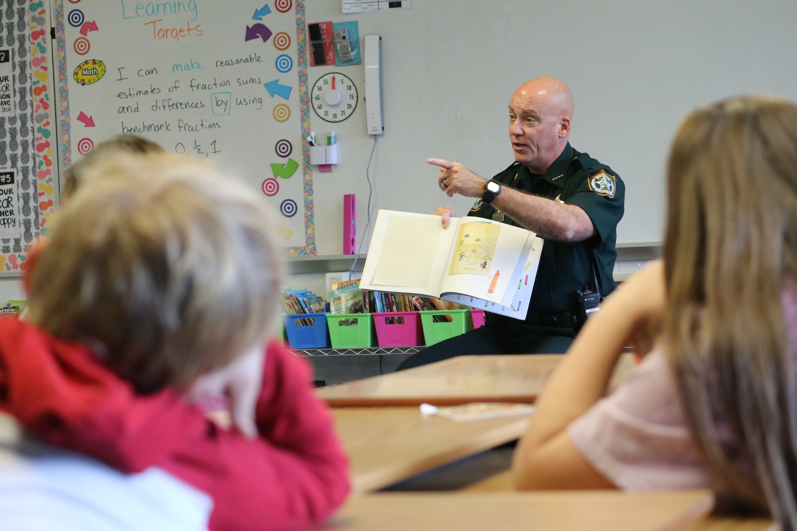 A police officer reading a book to elementary school children in a classroom, pointing at the book while explaining.