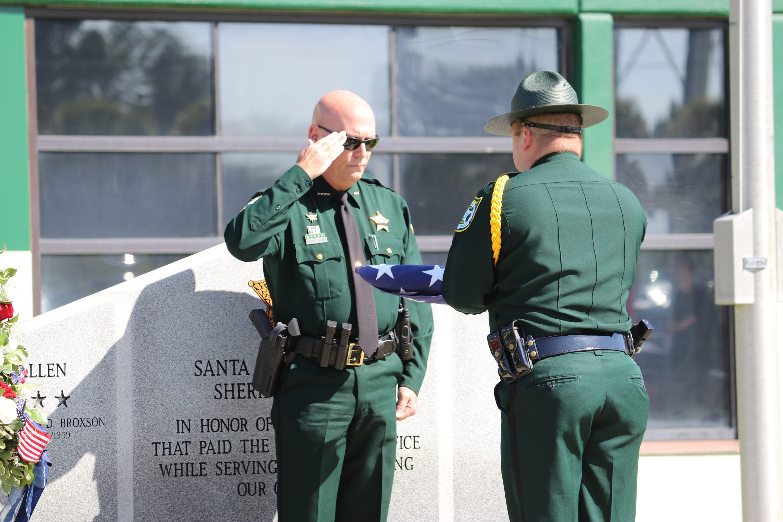 Two sheriff deputies saluting a memorial wreath at a monument during a commemorative ceremony.