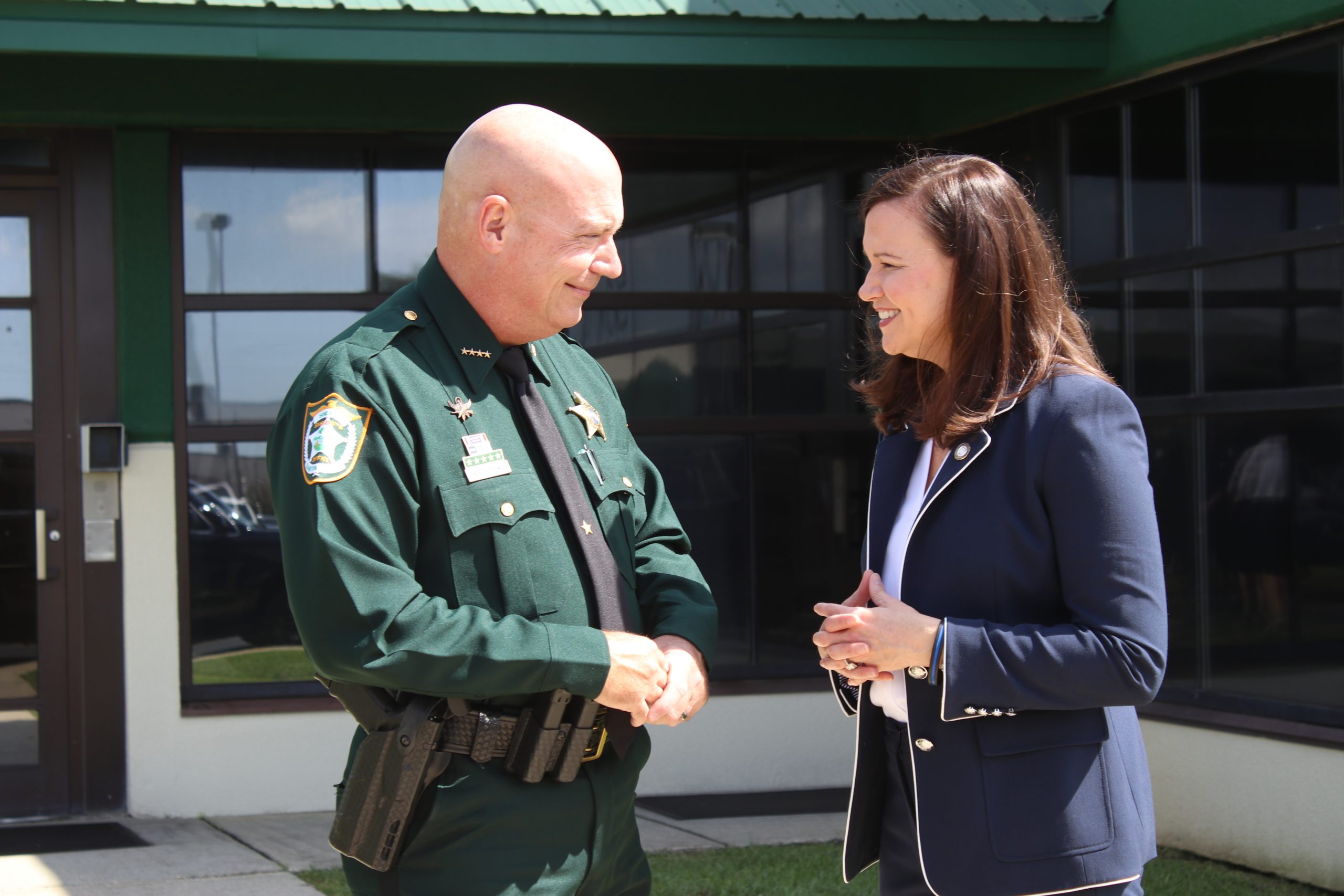 A sheriff in uniform shaking hands with a woman in a business suit outside a building on a sunny day.