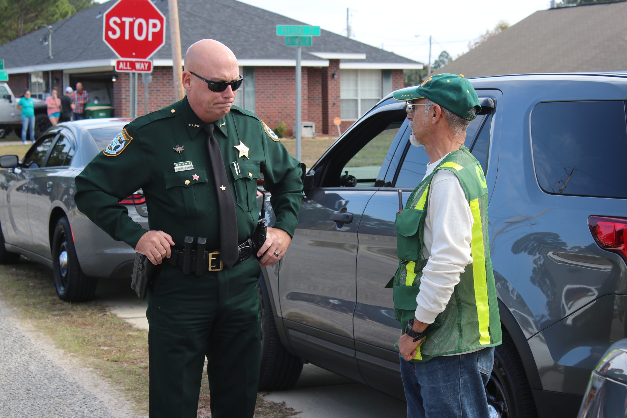 A sheriff talking to a man in a green vest next to a car, with a stop sign and other cars in the background.