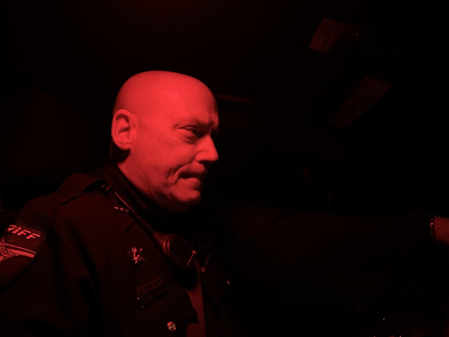 A bald police officer in a vehicle, illuminated by red light, looks to the side with a serious expression.