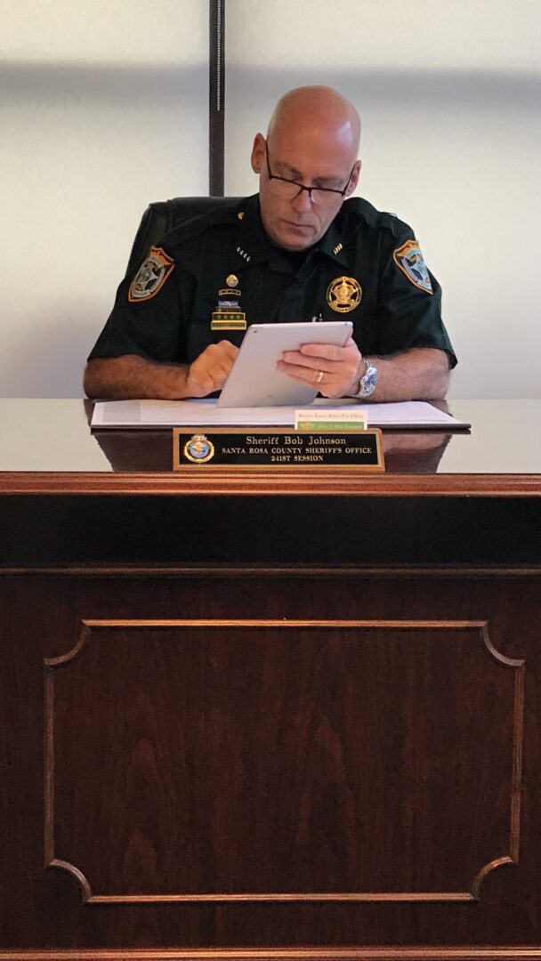 A sheriff in uniform, sitting at a desk, focused on writing in a notebook, with a nameplate reading "sheriff bob johnson" visible.