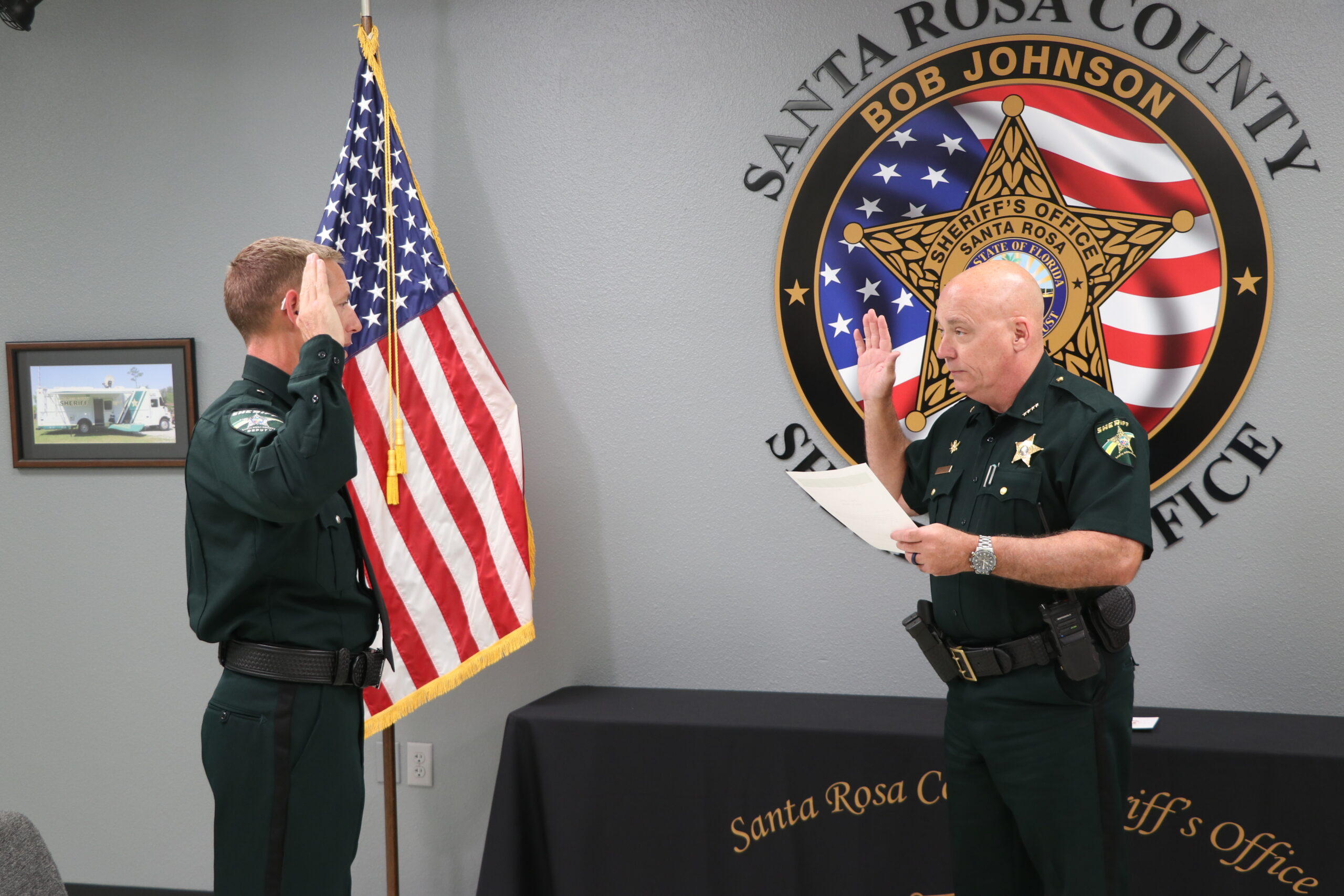 A sheriff's deputy is being sworn in by an officer in a room with american and state flags, and the department's seal visible.