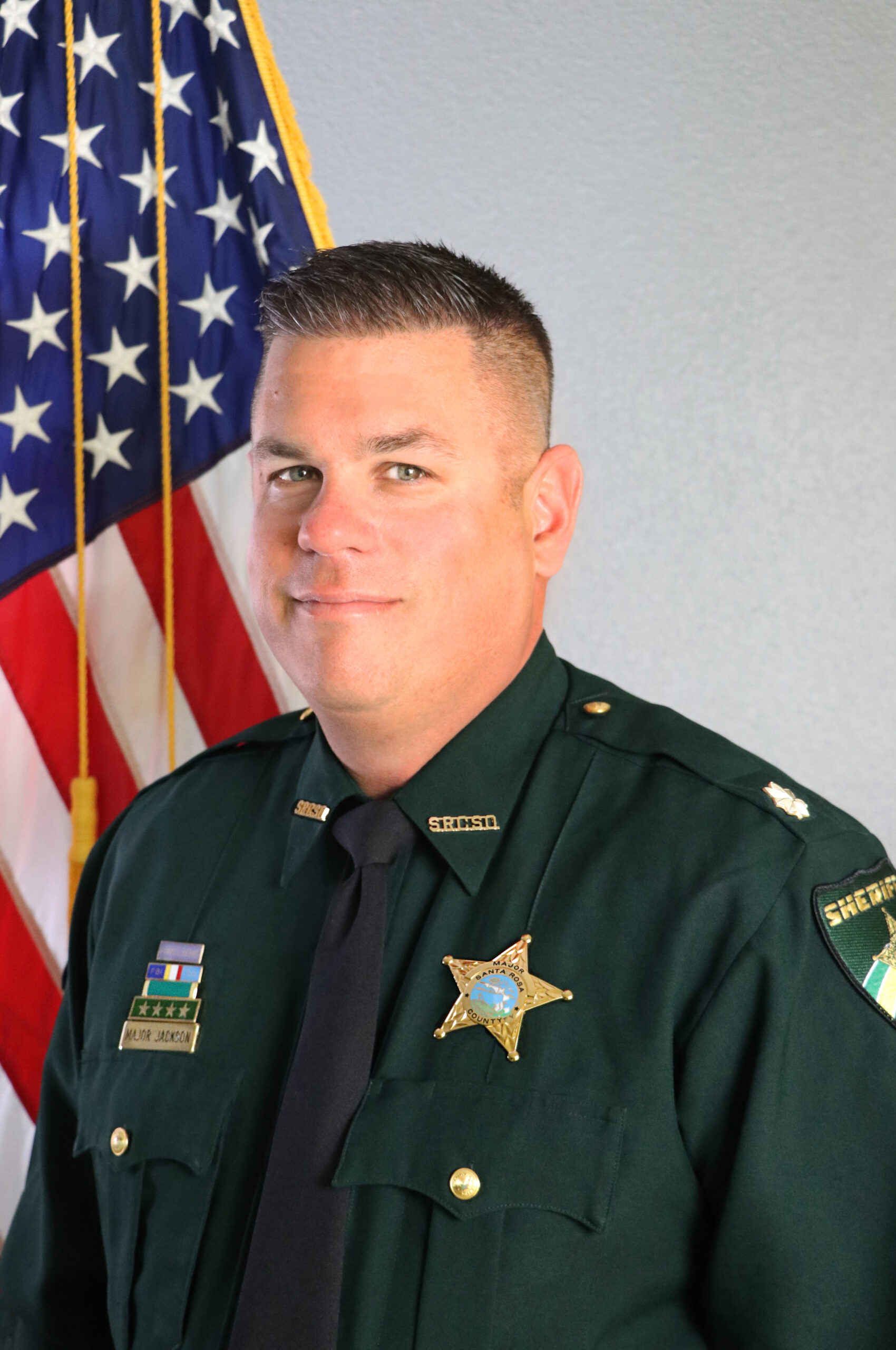 A sheriff's deputy in uniform, standing in front of an american flag, looking at the camera with a serious expression.