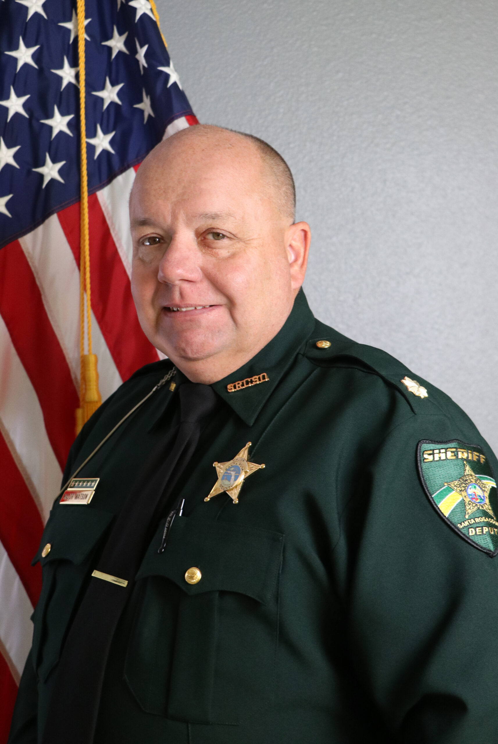 A smiling sheriff in uniform standing in front of an american flag.