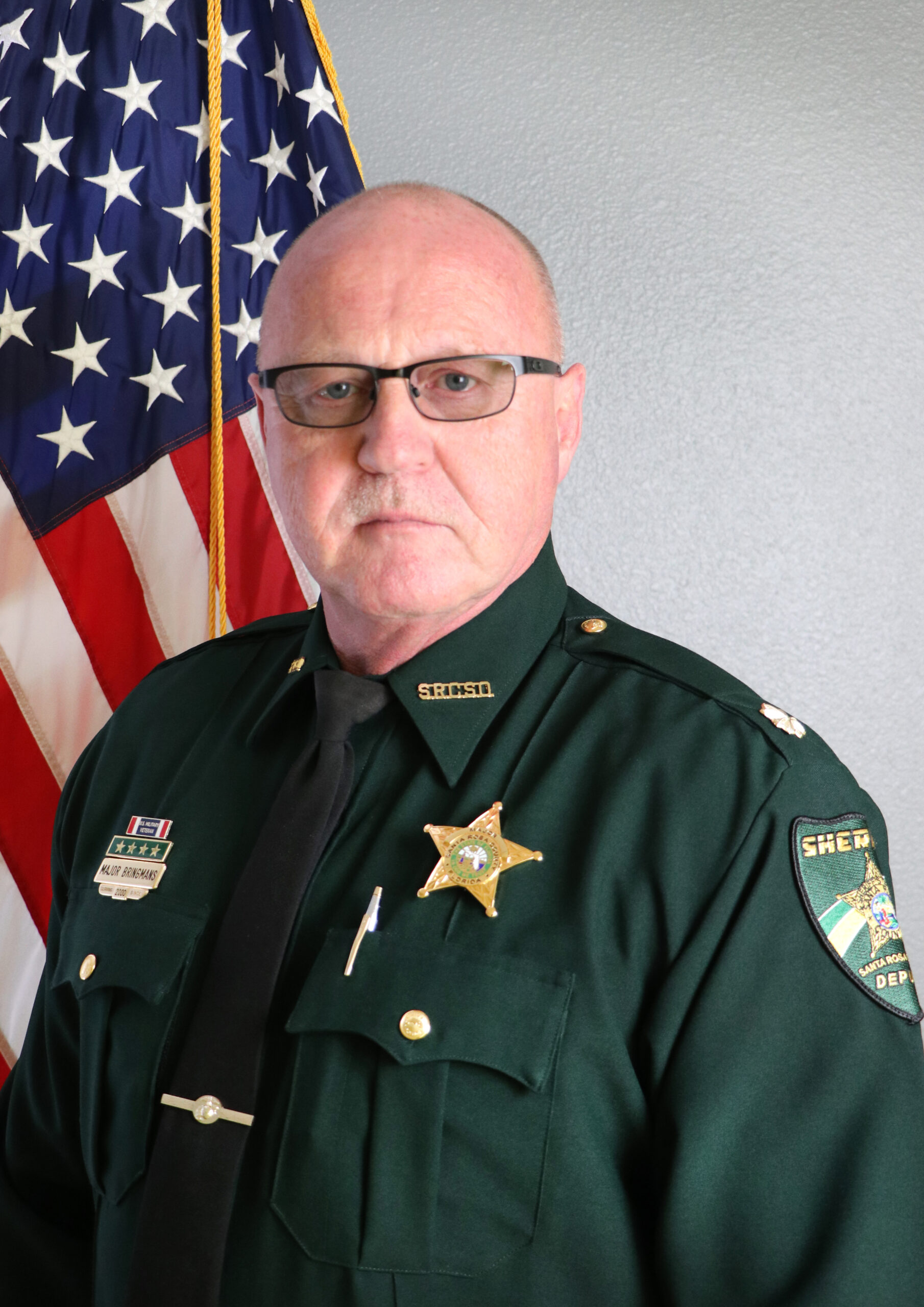 A middle-aged man in a sheriff's uniform stands in front of an american flag.