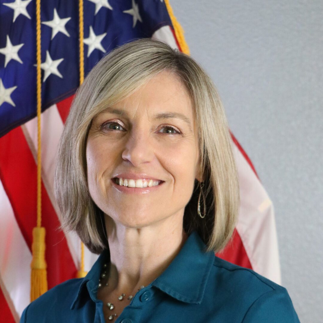 Portrait of a smiling woman in a teal shirt with short grey hair, standing in front of american flags.