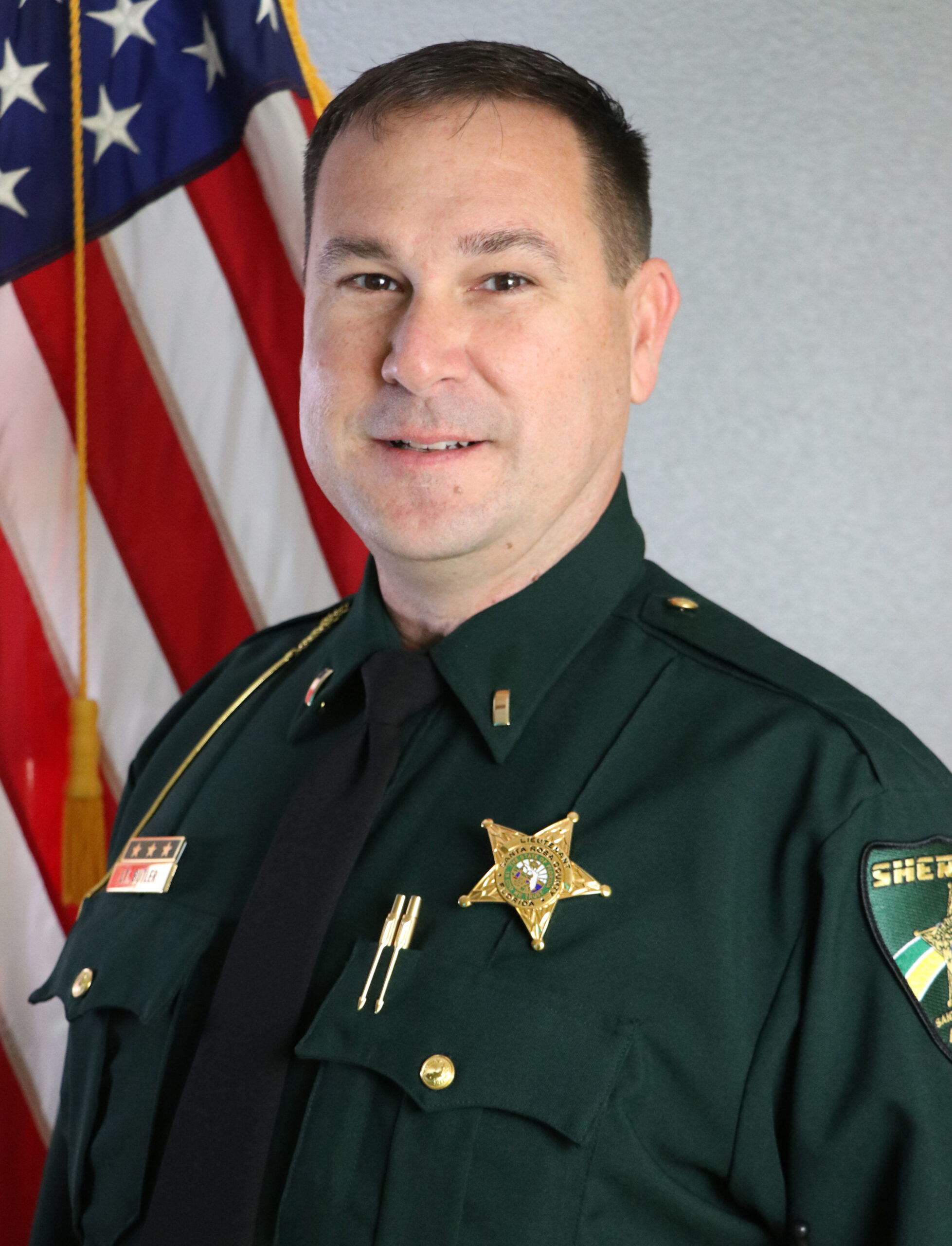 A portrait of a male sheriff in uniform with badges, standing in front of the american flag.