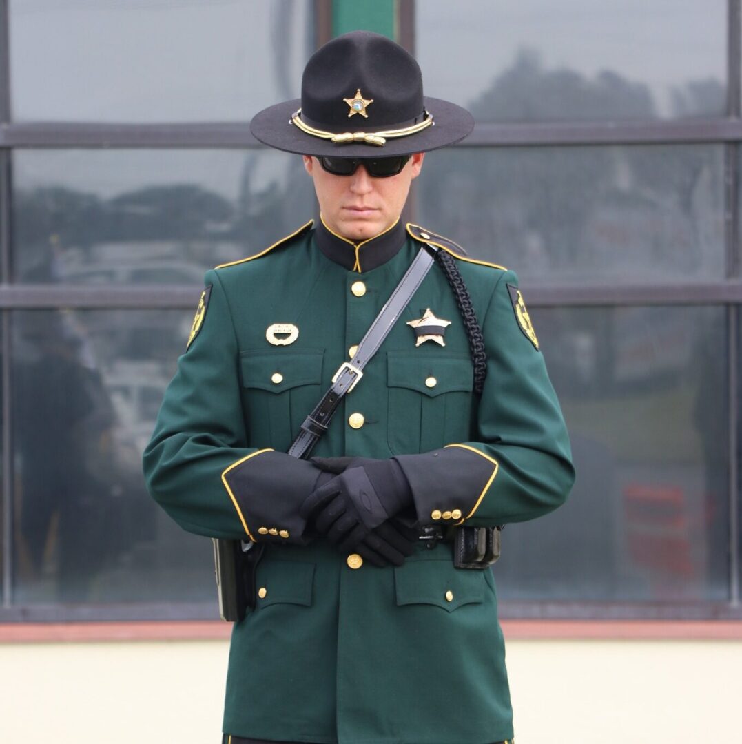 A sheriff wearing a green and black uniform with a star badge, sunglasses, and gloves, standing solemnly with arms crossed.
