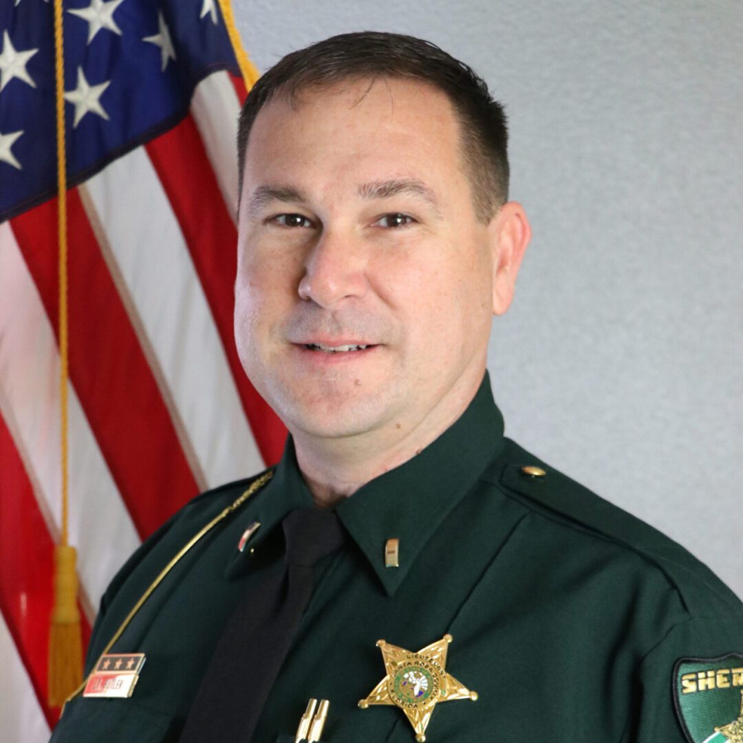 A portrait of a male sheriff in uniform with badges, standing in front of the american flag.