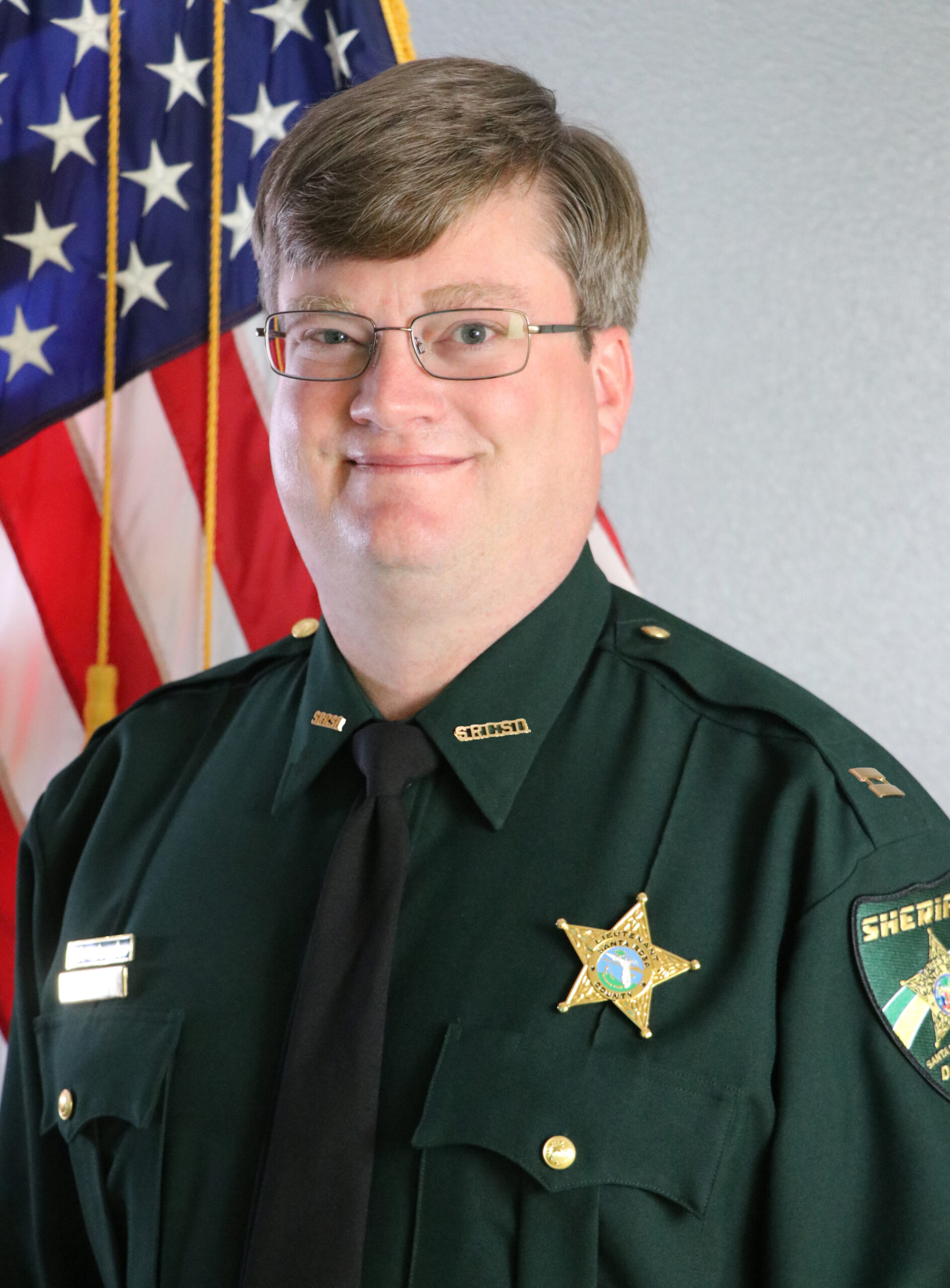 A sheriff in uniform smiling, standing in front of the american flag and another flag with a star emblem.