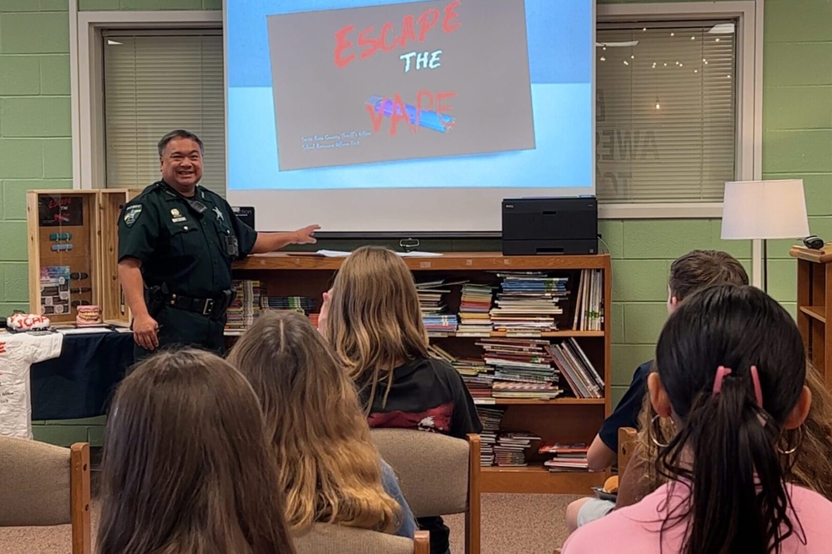 A police officer presenting at a school event, speaking to children who are seated, with a projection screen displaying "escape the vape" in the background.